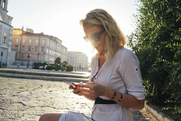 美丽而优雅的时尚白种人妇女用太阳镜使用手机 而坐在一个老城区广场的长凳上 — 图库照片