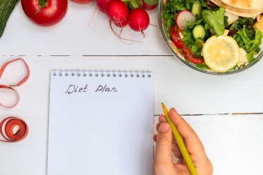 Bir kadın elinde kalem ve yazılar diyet planı beyaz ahşap masa üzerinde bir kase salata ve sebze ile