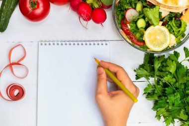 Beyaz boş not defteri ve sağlıklı salata ve taze sebzelerle beyaz ahşap masada elinde kalem tutan kadın manzarası. Sağlıklı diyet kavramı