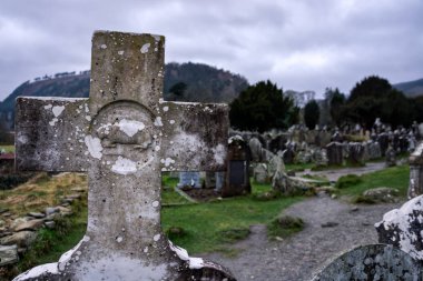 Arka planda mezarlık ve mezar taşları ile mezar taşı üzerinde eski katolik granit çapraz, Glendalough, İrlanda
