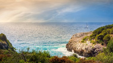 Kayalıklarla kıyı şeridi, turkuaz su ve yelkenli, gün doğumu, Mallorca, İspanya