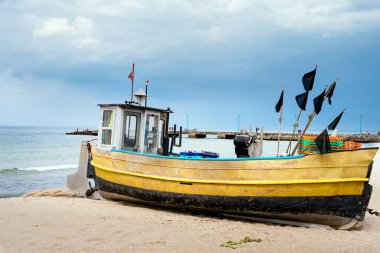 Arka planda mavi ve iskele olan kumlu sahilde paslı bir balıkçı teknesi.
