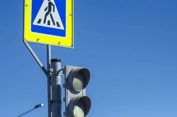 Traffic sign - a pedestrian crossing against the blue sky. A square blue-yellow crossing sign across the road. A white triangle road sign on a blue background.