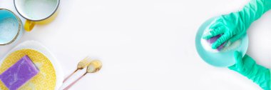 Pankart: Nane mavisi eldivenli eller mor süngerle mavi tabağı, tabakları, kupaları, çatalı ve kaşığı köpükle yıkıyor. Beyaz bir arkaplanda izole et. Temizlik konsepti. Metin için boşluk.