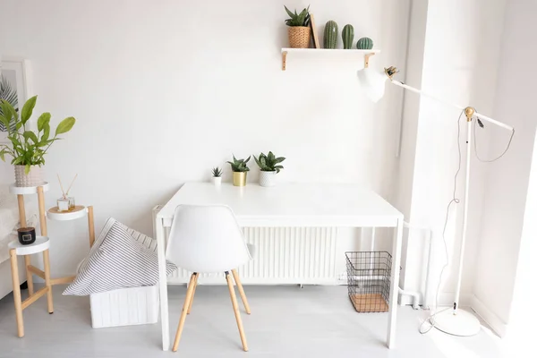 Dairede parlak bir çalışma alanı. Üç çiçekli beyaz çalışma masası, bir raf, bir lamba, bir sandalye, yastıklı bir sepet ve çiçekli ve güzel kokulu bir tezgah..