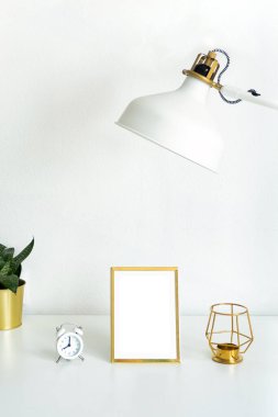 Beyaz masa, kapalı çiçek, beyaz çalar saat, altın şamdan ve beyaz lamba..