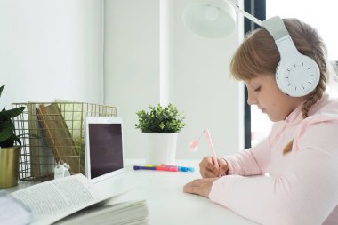 Genç kız elinde tabletle ve kulaklıkla bir masada oturuyor ve bir deftere yazıyor. uzaklık öğrenme kavramı.