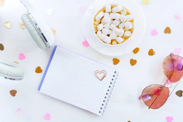 Beyaz kulaklıklar, kahve ve marşmelovlu bir fincan, konfeti kalpli beyaz bir masada bir not defteri ve bardaklar. yaşam tarzı, üst görünüm, düz görünüm