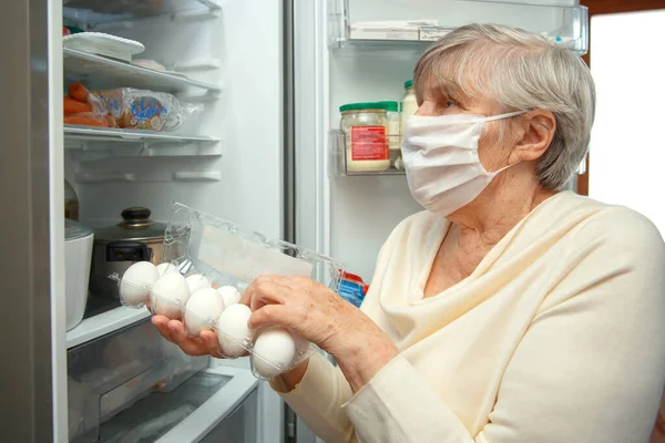 医療用マスクの高齢女性は パッケージから食品を取り 冷蔵庫に入れます 高齢者への食品配送 — ストック写真
