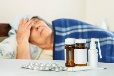 Yatakta yaşlı bir kadın, önünde masanın üstünde ilaçlar, masanın üstünde de ilaçlar var.