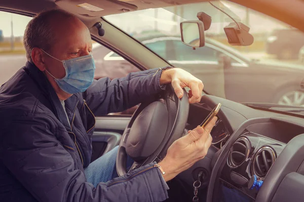 A man in a car with a phone in his hand and a protective mask against the spread of Coronavirus Covid-19