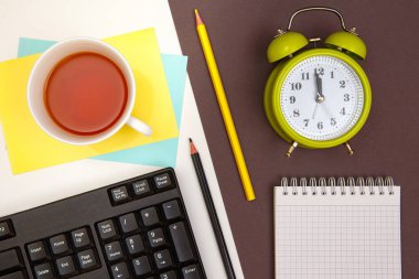 Not defteri, klavye, hesap makinesi ve kalemler, çalar saat ve çay fincanı. Beyaz ve koyu gri arkaplan.