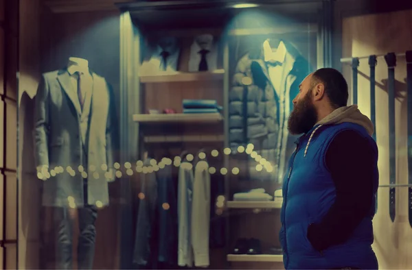 A bearded man in blue sporstwear looks to the shop window with fashionable business clothing on the manikin. Shirt and tie and grey men coat in the shop window.