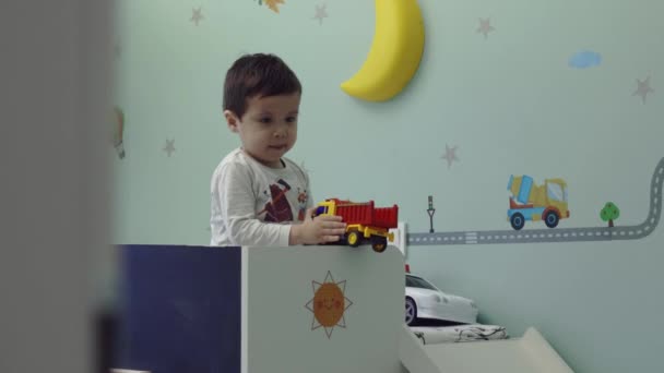 Bonito criança sentada na cama em seu quarto e brincando com brinquedos de caminhão, câmera no controle deslizante — Vídeo de Stock