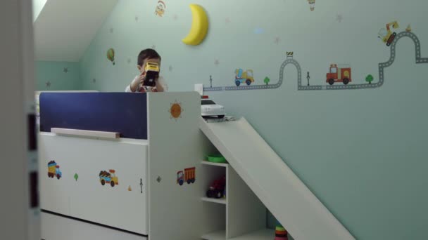Criança bonito sentado em sua cama na sala de crianças e brincando com brinquedos de caminhão e fazer rosto surpreso, câmera no controle deslizante — Vídeo de Stock