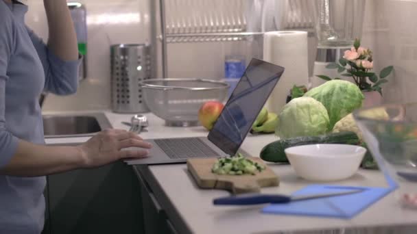 Chiudi giovane donna in cerca di un fresco insalata recept nel computer portatile in cucina. Telecamera sul cursore. Preparazione di insalata fresca con cipolla, cetriolo e ravanelli . — Video Stock