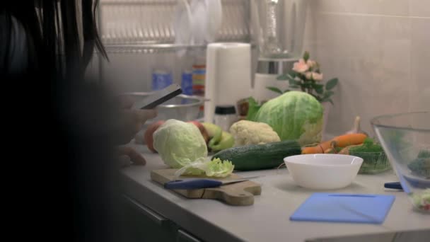 Bella giovane donna che prepara l'insalata in cucina mentre tiene un telefono cellulare. Girato sul cursore — Video Stock