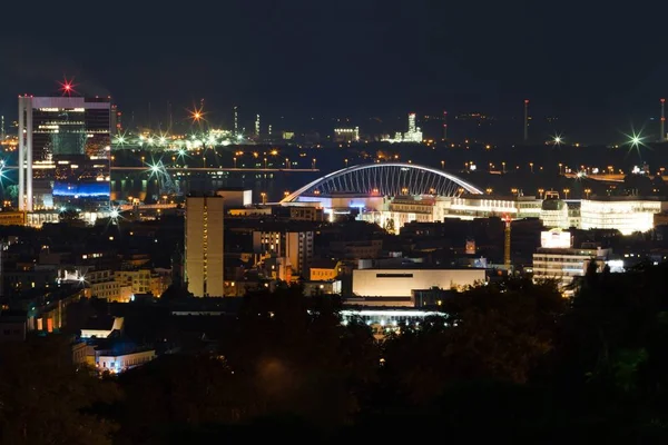 Natt syn på staden. Röda lampor, byggnader, bro och fabriken. — Stockfoto