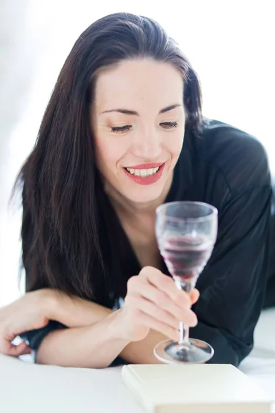 Hübsche Frau in Schwarz liegt auf dem Bett und schaut in ein Glas Wein. — Stockfoto