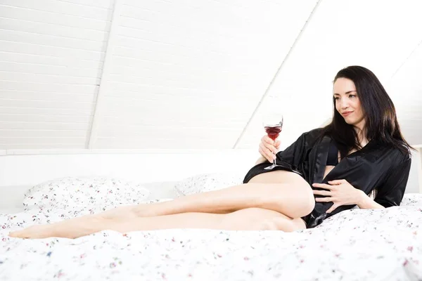 Piękna młoda kobieta w szlafrok z kieliszek czerwonego wina w sypialni. — Zdjęcie stockowe