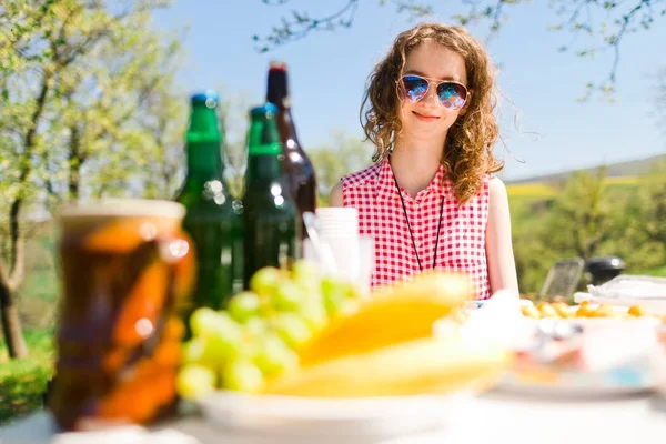 Adolescente di età compresa tra ragazza in camicia rossa a scacchi seduta al tavolo sulla festa in giardino cibo e bottiglie sul tavolo . — Foto Stock