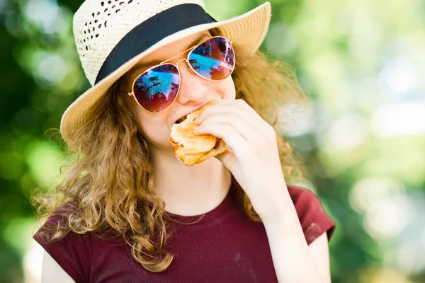 Güneş gözlüklü şapkalı bir kız hamburgeri ısırdı.