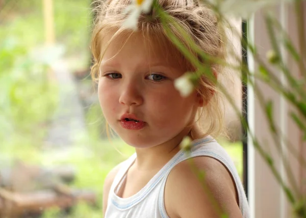 portrait of a little girl in a summer white t-shirt with blond hair looking out from behind the stalks of daisies