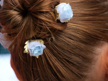 Sarı saçlar düzgünce taranır, at kuyruğunda yığılır, bükülmüş ve saplanmış saç tokaları mavi yapay güllerle süslenir.