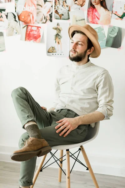 Homem elegante e bonito em um chapéu sentado na cadeira perto da parede branca — Fotografia de Stock