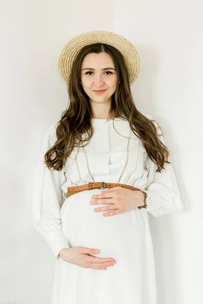 Молода красива вагітна жінка в капелюсі стоїть біля білої стіни — стокове фото