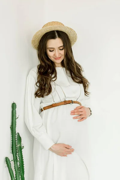Молода красива вагітна жінка в капелюсі стоїть біля білої стіни — стокове фото