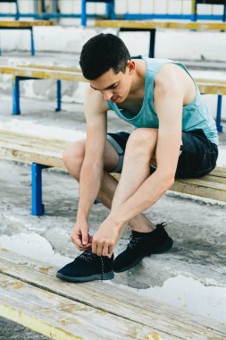 Bir bankta oturan ve ayakkabı bağcıklarını temiz havaya bağlayan genç bir adam.