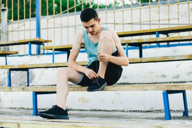 Bir bankta oturan ve ayakkabı bağcıklarını temiz havaya bağlayan genç bir adam.