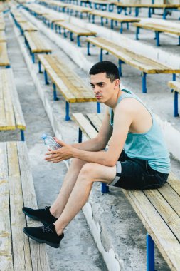 Bir bankta oturan ve ayakkabı bağcıklarını temiz havaya bağlayan genç bir adam.