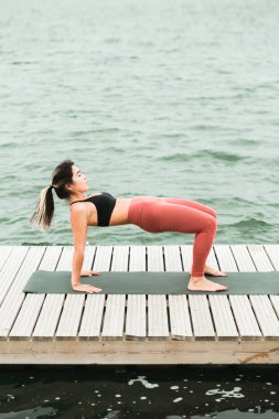 Asyalı genç kız göl kenarında iskelede açık havada yoga yapıyor.