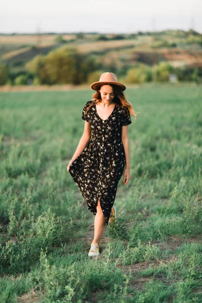 Молода, ніжна дівчина в капелюсі ходить по зеленій галявині в промені заходу сонця — стокове фото