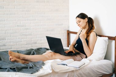 Avrupalı bir kadın, dizüstü bilgisayar kullanarak, yatağında internetten bilgi öğrenirken, virüs karantinadayken günlüğüne notlar yazıyor. Rahat bir iş yeri, e-öğrenim konsepti