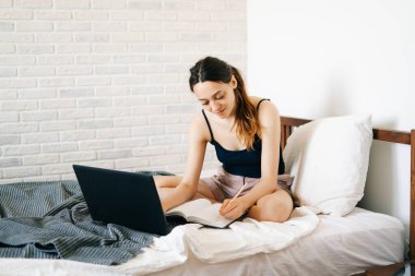 Avrupalı bir kadın, dizüstü bilgisayar kullanarak, yatağında internetten bilgi öğrenirken, virüs karantinadayken günlüğüne notlar yazıyor. Rahat bir iş yeri, e-öğrenim konsepti