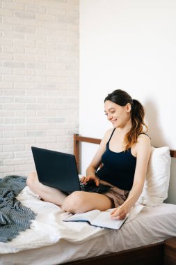 Avrupalı bir kadın, dizüstü bilgisayar kullanarak, yatağında internetten bilgi öğrenirken, virüs karantinadayken günlüğüne notlar yazıyor. Rahat bir iş yeri, e-öğrenim konsepti