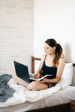 Avrupalı bir kadın, dizüstü bilgisayar kullanarak, yatağında internetten bilgi öğrenirken, virüs karantinadayken günlüğüne notlar yazıyor. Rahat bir iş yeri, e-öğrenim konsepti