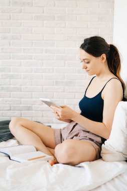 Tablet kullanan genç Avrupalı bir kadın evinden uzakta, yatağında çalışır ve kendi kendini izole ederken günlüğüne notlar alır. Rahat bir iş yeri, e-öğrenim konsepti. Uzak çalışma kavramı. Serbest.