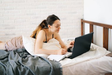 Avrupalı bir kadın, dizüstü bilgisayar kullanarak, yatağında internetten bilgi öğrenirken, virüs karantinadayken günlüğüne notlar yazıyor. Rahat bir iş yeri, e-öğrenim konsepti