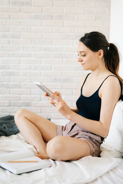 young European woman using a tablet works remotely from home,in her bed and makes notes in her diary,while self-isolating.cosy working place,the concept of e-learning. concept of remote work.Freelance