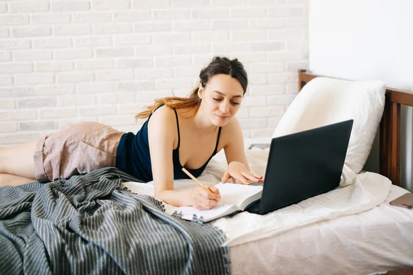 Avrupalı bir kadın, dizüstü bilgisayar kullanarak, yatağında internetten bilgi öğrenirken, virüs karantinadayken günlüğüne notlar yazıyor. Rahat bir iş yeri, e-öğrenim konsepti
