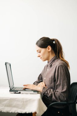 Bilgisayarında virüs karantinası sırasında evden çalışan genç bir Avrupalı kadın var. Kız minimalist bir iç mekanda beyaz bir masada oturuyor. Rahat bir iş yeri, uzaktan çalışma konsepti.,