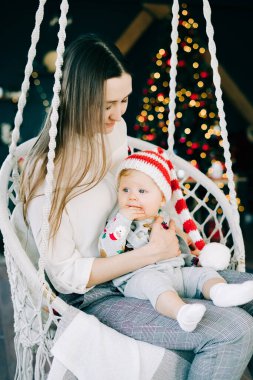 Çocuğun ilk Noel 'i. Anne, parlak bir Noel ağacı konseptinin arka planında hasır bir salıncakta oturan Noel Baba şapkalı küçük bir çocuğu tutuyor..