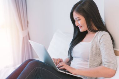 Dizüstü bilgisayarda çalışan genç bir kadın evde yatağında oturuyor.