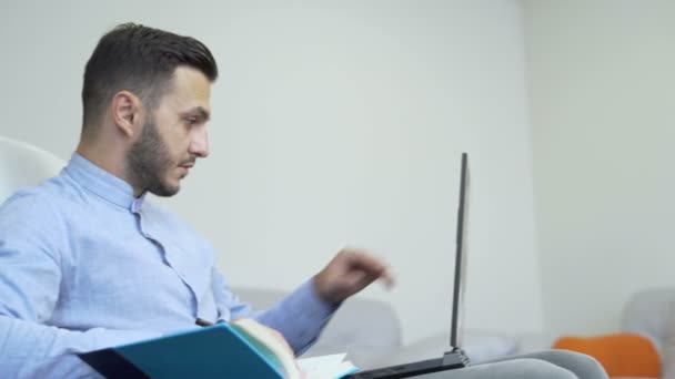 Joven hombre caucásico en camisa azul claro trabajando con portátil y tomando notas — Vídeo de stock