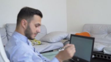 Ciddi genç işadamı ev ofisinde dizüstü bilgisayar üzerinde çalışan ve kalem ile notlar alarak, yakından bakmak