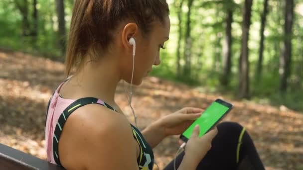 Kulaklıklı, telefondan şarkı seçen ve ormanda sabah egzersizi yapmaya hazırlanan muhteşem bir kadının yan görüntüsü. — Stok video